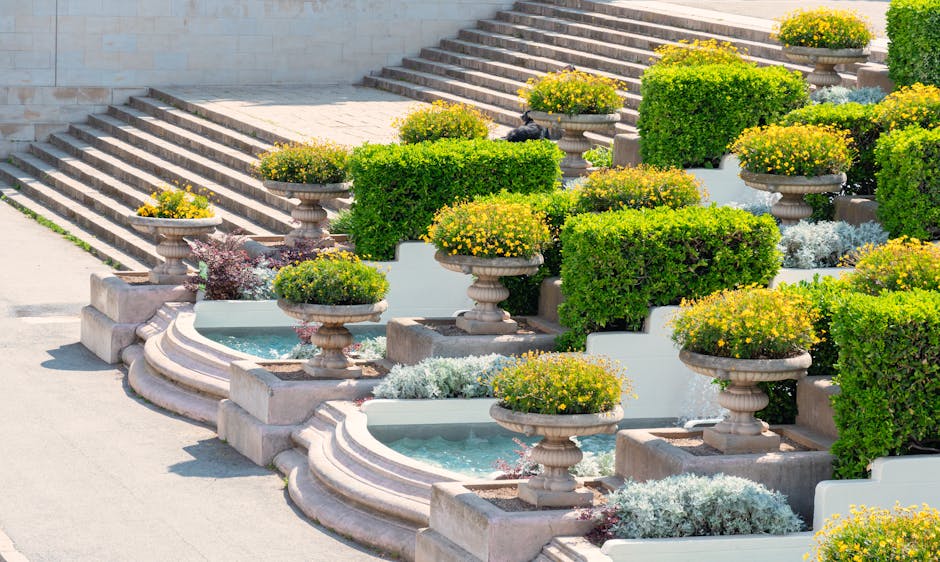 A tiered outdoor garden feature with wide stone steps leading up to a terrace, adorned with symmetrical plant arrangements in classical urns. The urns contain vibrant yellow flowering plants and are p
