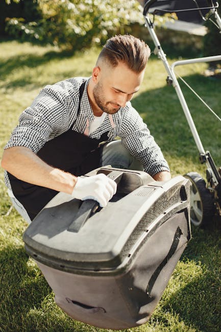 A man with styled brown hair and a trimmed beard is kneeling on a lush green lawn, carefully opening the lid of a grey outdoor garden waste bin. He is wearing a checkered shirt, a black gardener's apr
